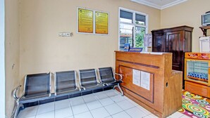 A hotel reception area with a wooden desk, a receptionist sign, and a window. There are four metal chairs lined up against the wall. - OYO 92287 Hotel Aji Mantoro (Belik)
