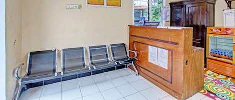 Zona de recepción de hotel con un mostrador de madera, un letrero de recepcionista y una ventana. Hay cuatro sillas metálicas alineadas contra la pared.