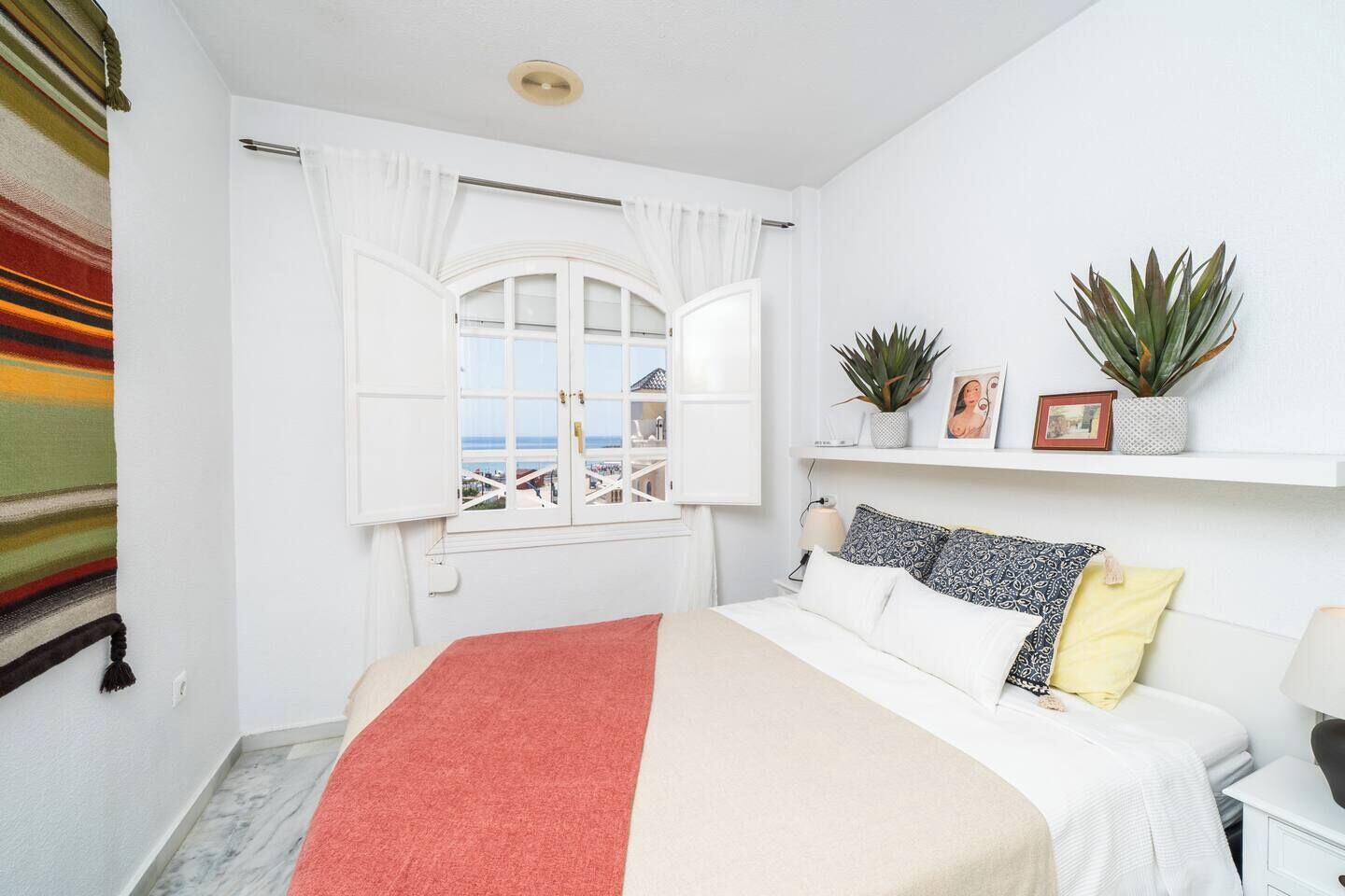 Une chambre à coucher avec un lit, une fenêtre avec des rideaux, une plante sur une étagère et une vue sur la mer.