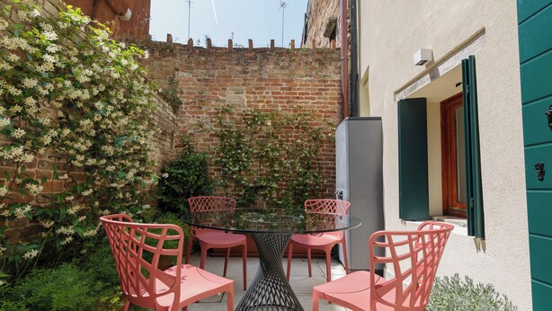 A glass-top table with four pink chairs in a courtyard with a brick wall and potted plants. - Venice • Private Court / Peggy Cozy Apartment (Venezia)