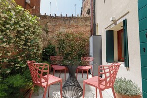 Une table à plateau en verre, entourée de quatre chaises roses, se trouve dans une cour bordée d’un mur de briques et agrémentée de plantes en pot.