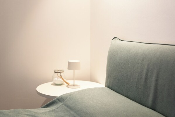 Un coin de pièce avec un fauteuil vert recouvert de tissu, une petite table d’appoint blanche sur laquelle se trouve une lampe, et un bocal en verre avec un couvercle en métal.