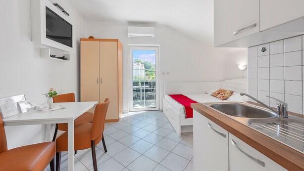 Une cuisine compacte avec un évier, un coin repas comprenant une table et des chaises, un téléviseur fixé au mur et une porte coulissante donnant sur un espace extérieur.