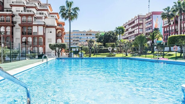 A swimming pool with a clear blue water surface, surrounded by a fence, and adjacent to a building with balconies and a garden area. - Serene Seaside Stay | Pool, Garden & Balcony (Torrevieja)
