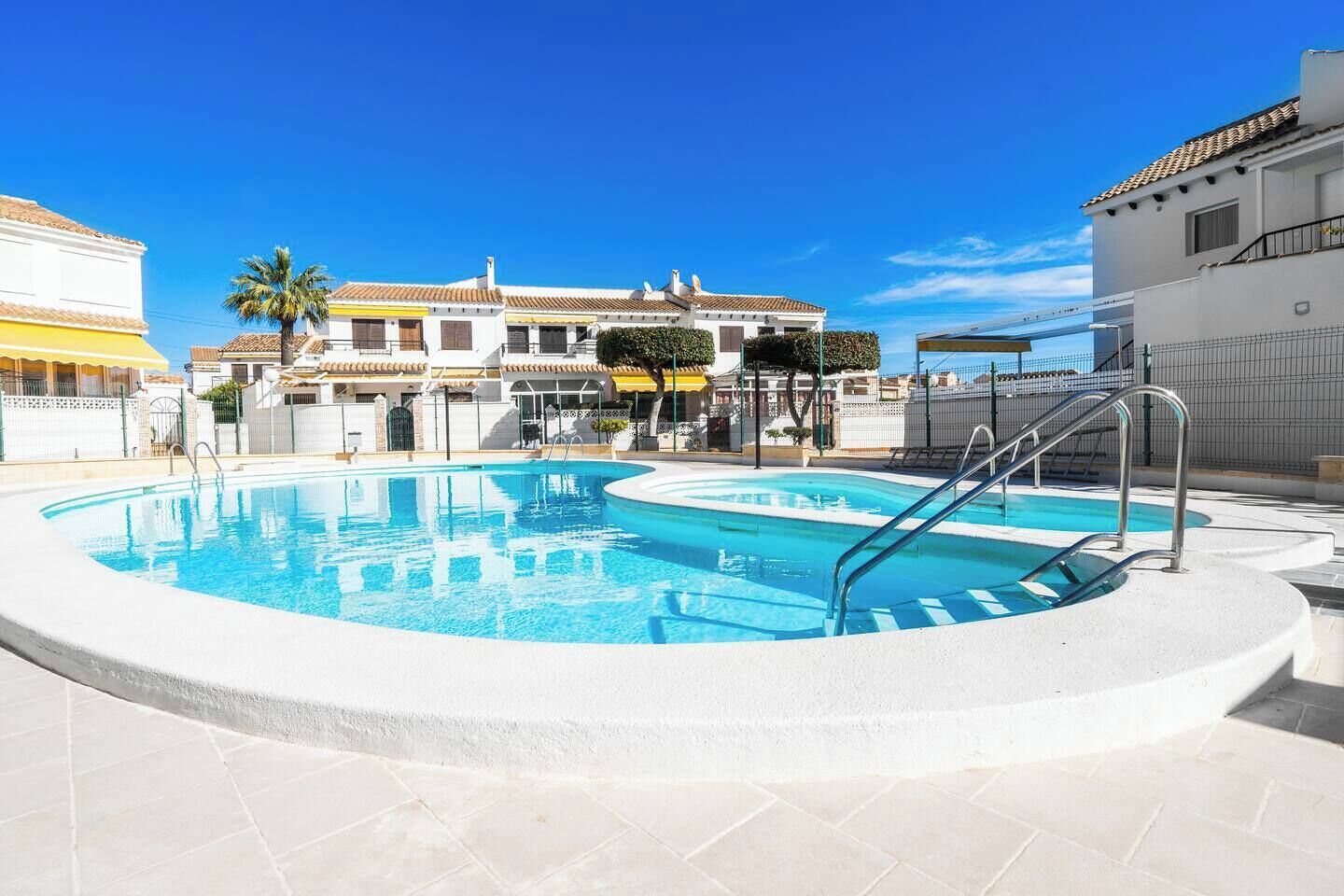 A swimming pool with a surrounding deck, a palm tree, and a building with a tiled roof in the background.