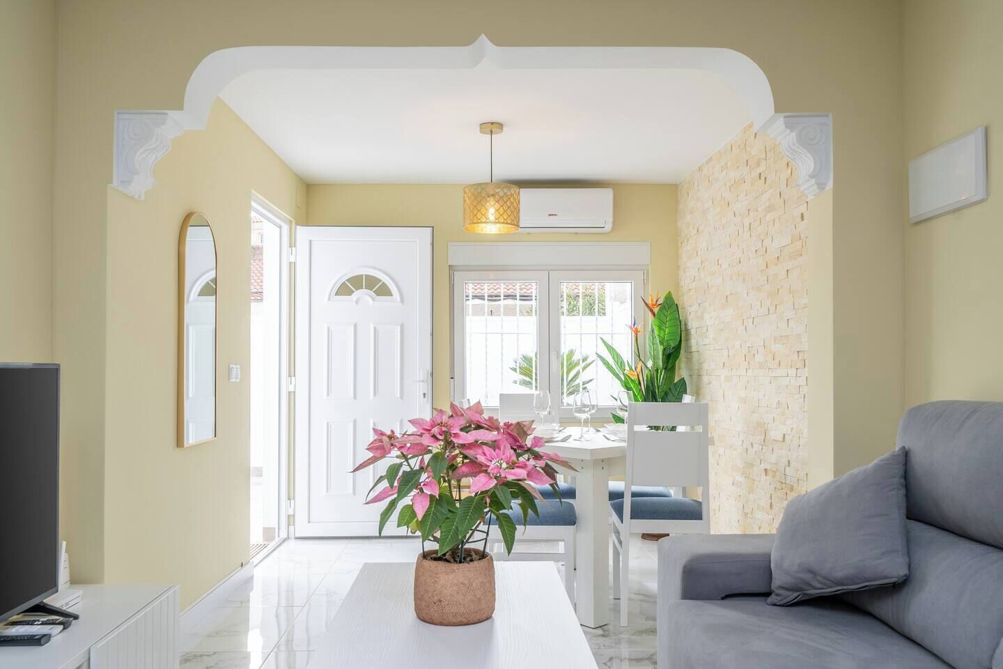 Una sala de estar con un sofá blanco, una mesa de centro con una planta en maceta, un televisor y un comedor con una mesa y sillas blancas.