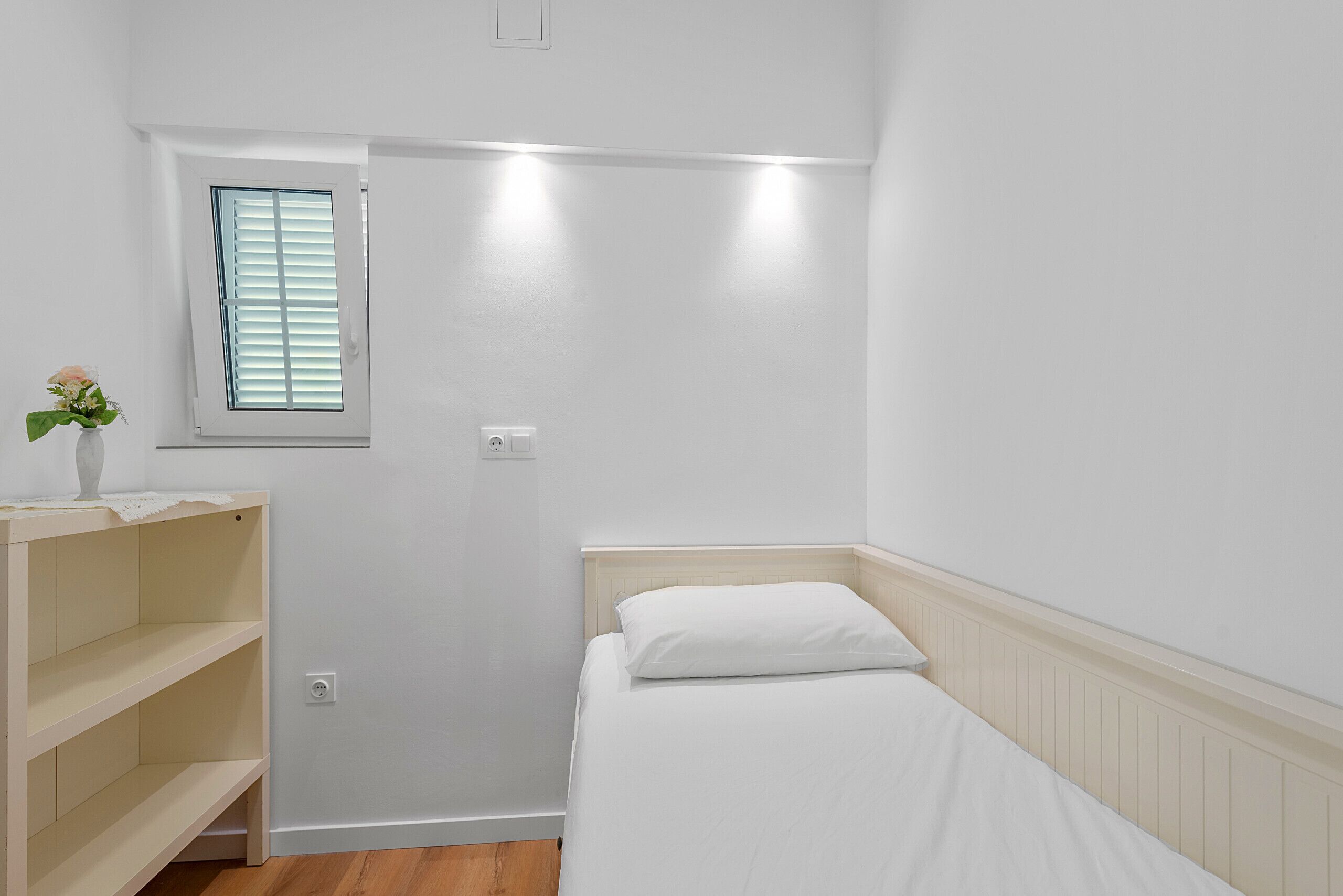 A small, clean room with a single bed, a window with blinds, a shelf unit, and a wall-mounted light fixture.