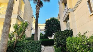 A narrow pathway flanked by lush greenery and tall palm trees, leading to a building with a yellow facade and a balcony.