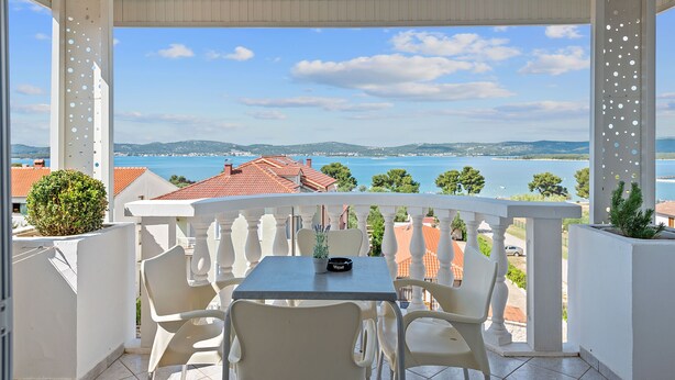 A balcony with a table and chairs, overlooking a coastal view with houses and the sea.