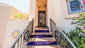 Un escalier menant à une porte ornée d’une rampe métallique, d’une décoration en forme de soleil et d’une fenêtre avec un grillage décoratif.