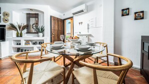 Un coin repas avec une table en verre entourée de chaises en osier, un mur blanc agrémenté d’objets décoratifs et une vue sur l’extérieur par une porte ouverte.