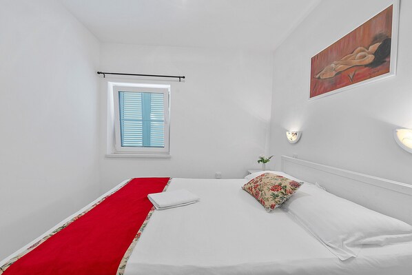 Una cama bien tendida con sábanas blancas y un corredor rojo, una ventana con contraventanas azules, una lámpara fijada a la pared y un cuadro.