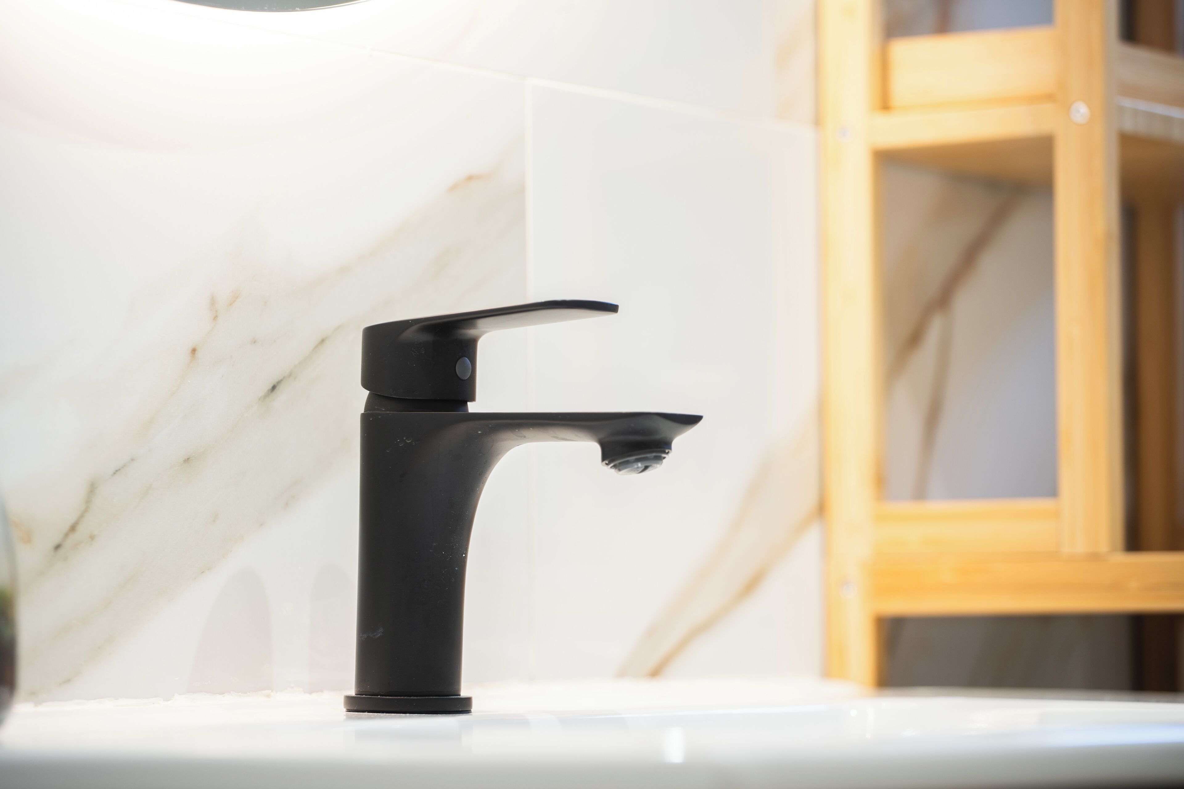 A modern bathroom with a black faucet on a white countertop, marble tiles, and a wooden cabinet in the background.