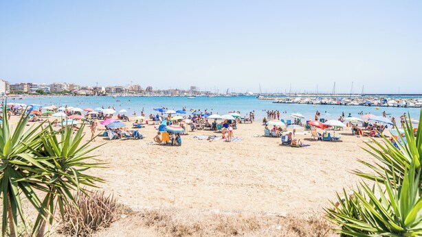 Une plage avec de nombreux parasols et beaucoup de monde, un port de plaisance avec des bateaux, et des bâtiments en arrière-plan.