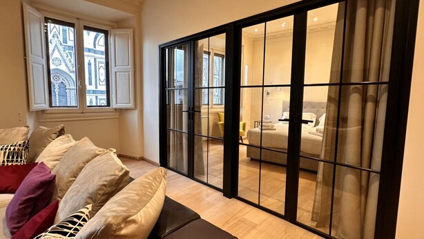 Een moderne woonkamer met een bank, een grote schuifdeur naar een slaapkamer, en een raam met decoratieve luiken.