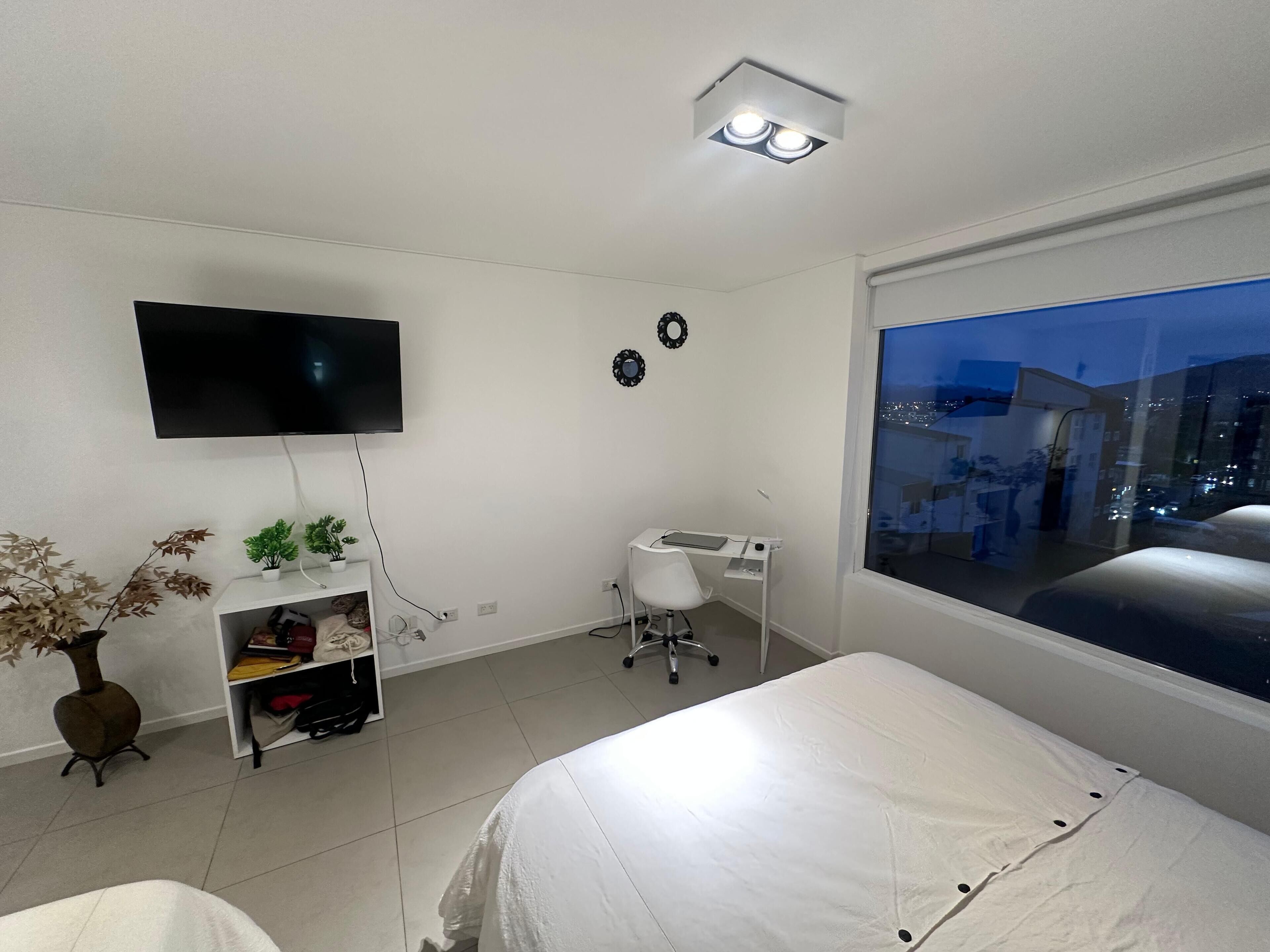 A bedroom with a bed, a desk, a chair, a TV, and a window with a city view.