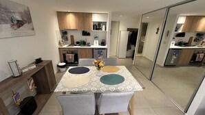 Una cocina moderna con armarios de madera, horno empotrado y refrigerador. Hay una zona de comedor con mesa y sillas, y un estante con objetos decorativos.