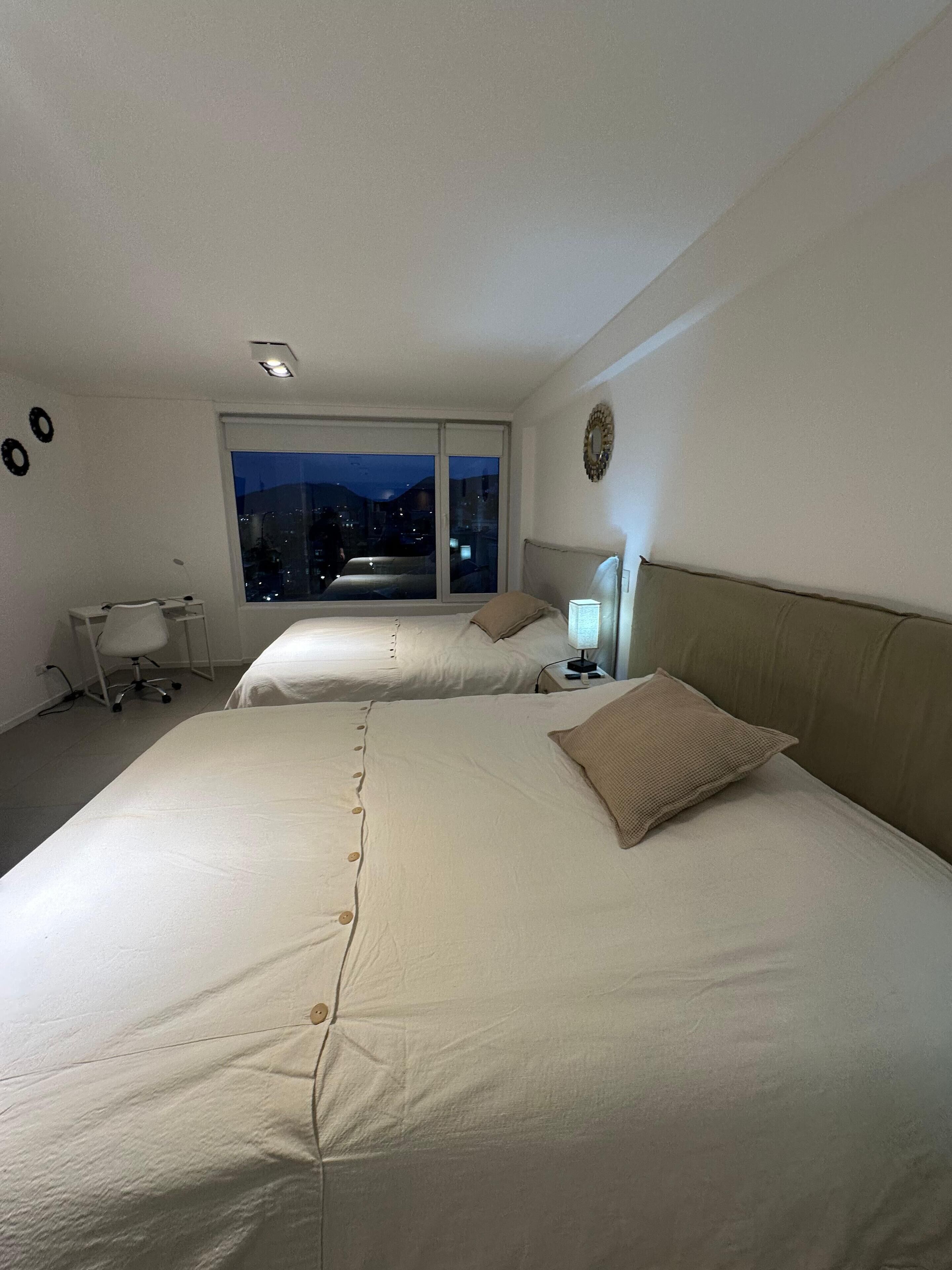 Habitación de hotel con dos camas, un escritorio y una silla. Se ve la ciudad iluminada desde la ventana.