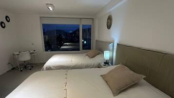 Habitación de hotel con dos camas, un escritorio y una silla. Se ve la ciudad iluminada desde la ventana.