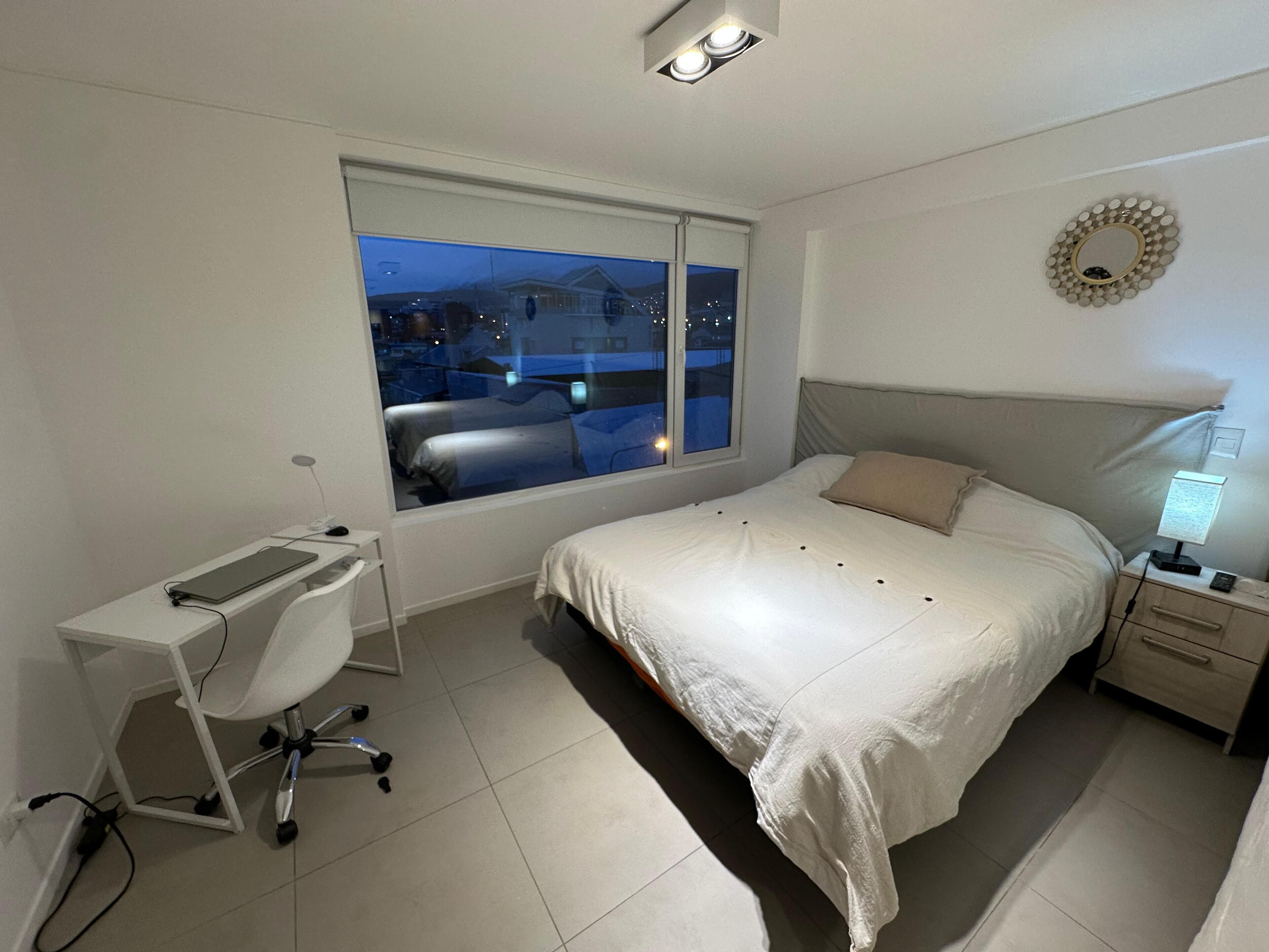Ein Schlafzimmer mit großem Fenster, einem Schreibtisch, einem Bett und einem Nachttisch.