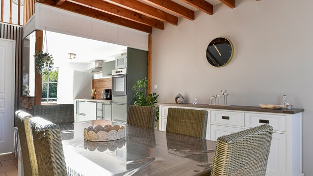 A dining area with a glass-top table, wicker chairs, a kitchen in the background, and a clock on the wall. - Cottage 'La Mottelette' with Private Terrace and Garden (Forest-Montiers)