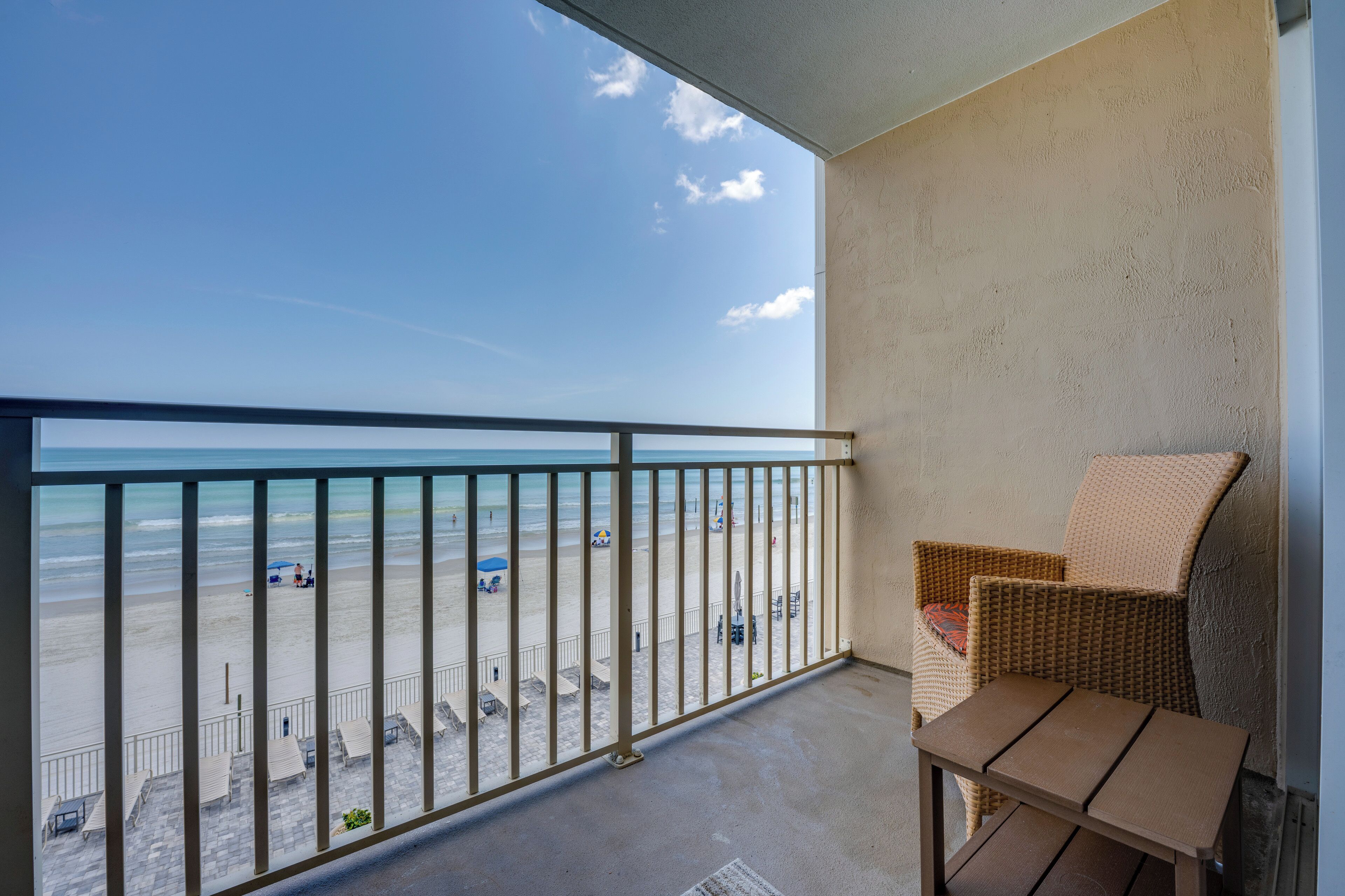 A balcony with a view of the beach, a wicker chair, and a small table.