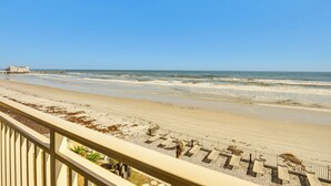 A beach view from a balcony with a railing, a pier in the distance, and a sandy beach with some vegetation. - Oceanfront Gem - Walk to Daytona Beach Pier! (Daytona Beach)