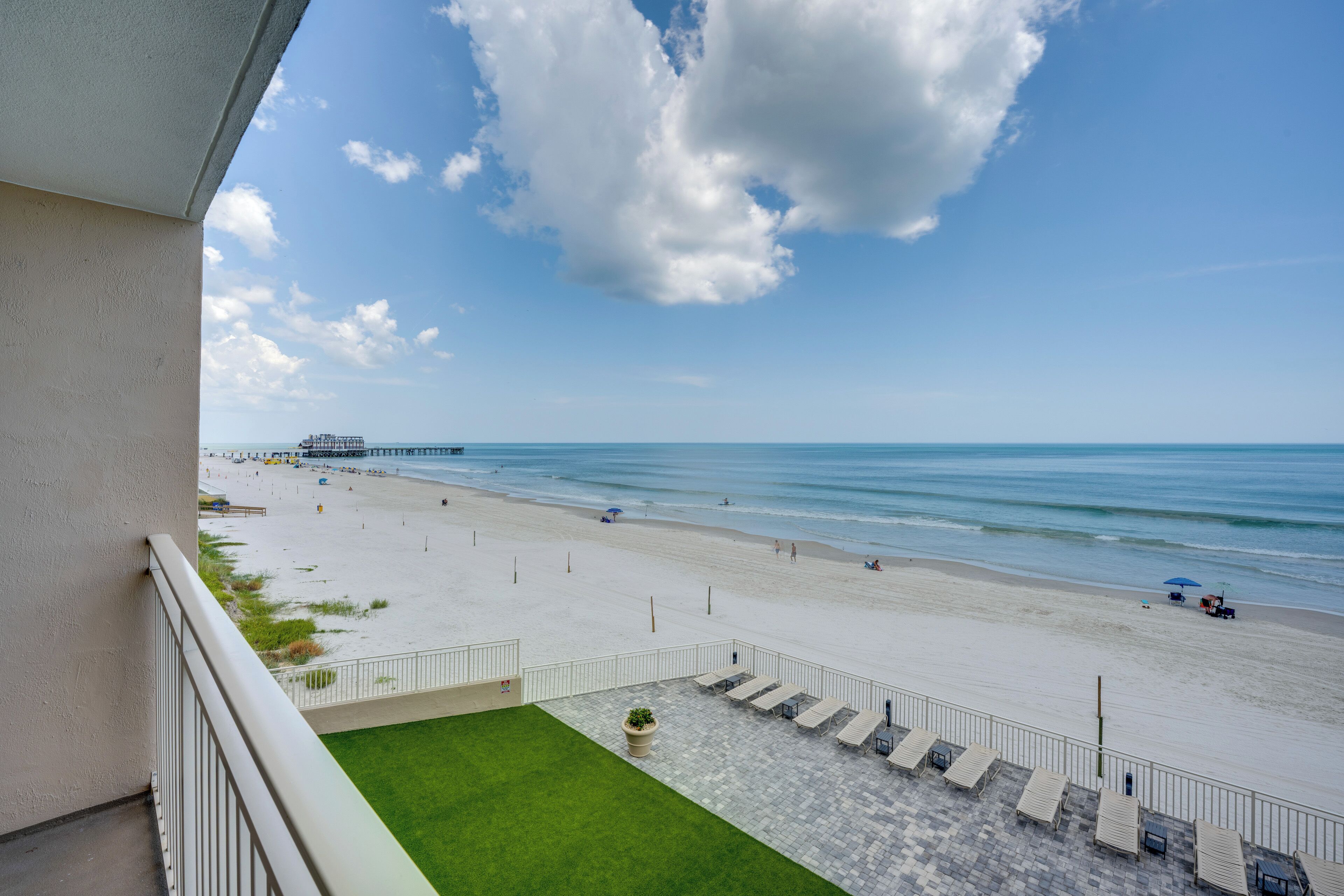 A beach view from a balcony with a clear sky, a sandy beach, and a pier in the distance.