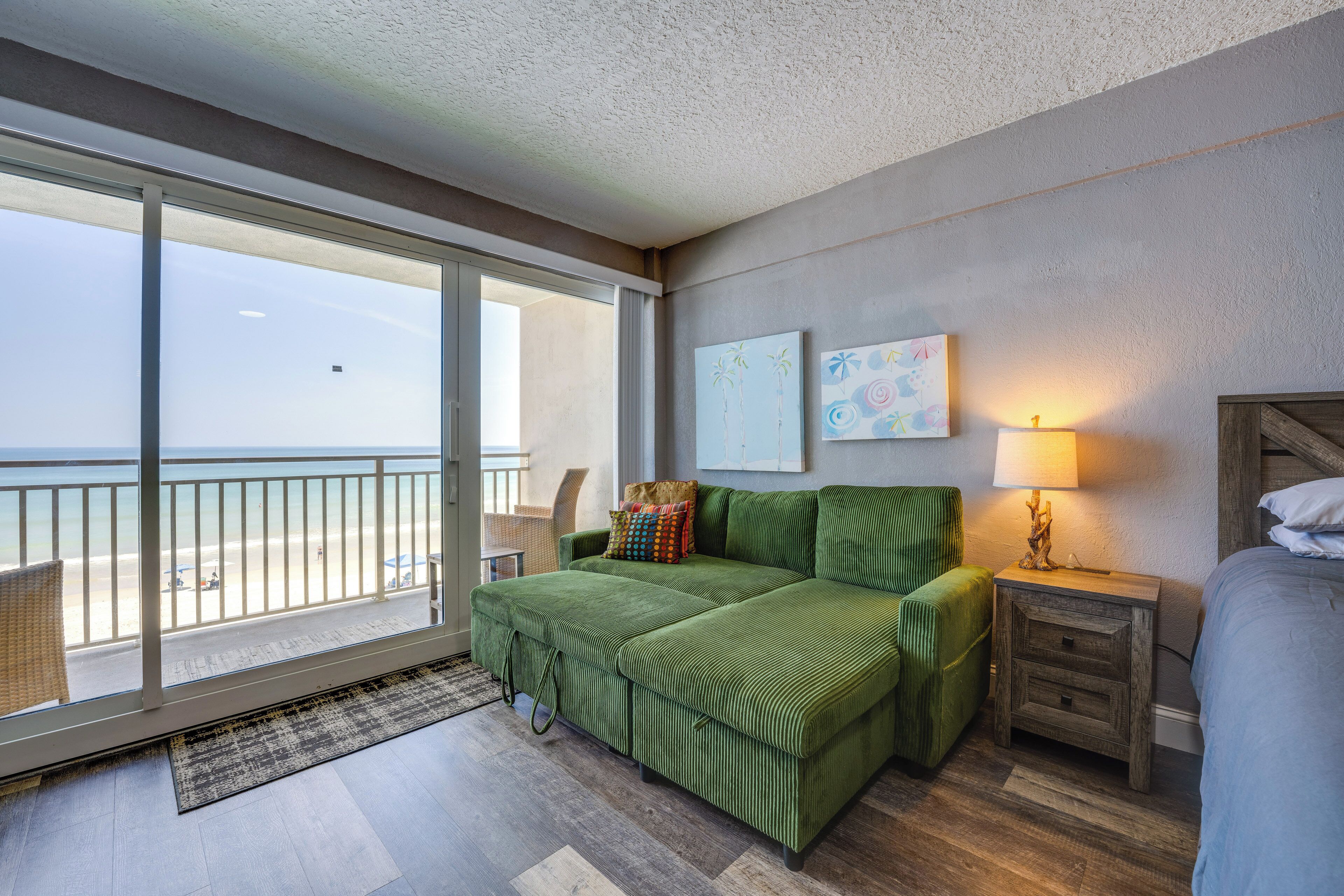 Une pièce avec un canapé vert, une table de chevet en bois, une lampe et une vue sur la plage, visible à travers une grande fenêtre.