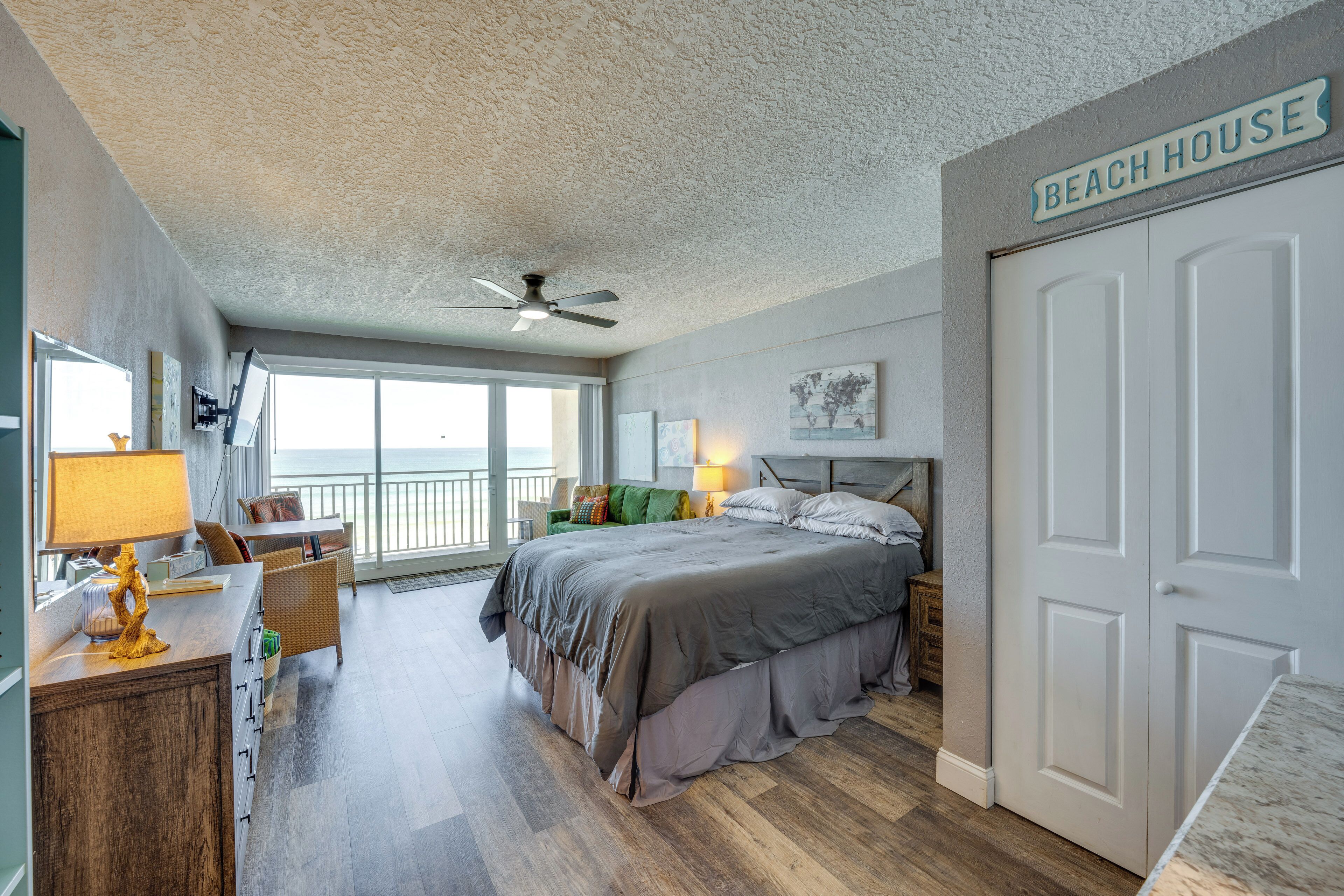 Una camera da letto con un letto, una scrivania, una sedia, una lampada, una ventola a soffitto, una finestra e una porta con la scritta 'BEACH HOUSE'.