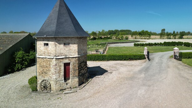 Une tour en pierre surmontée d’un toit conique, une allée gravillonnée et un champ clôturé.
