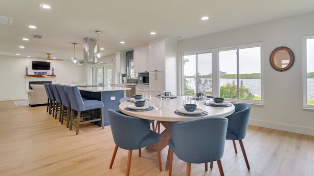 A modern kitchen with white cabinets, a dining area with a round table and blue chairs, and a view of a lake through large windows. - Riverfront Basecamp to Boat Fish Chase Sunsets (Ruskin)