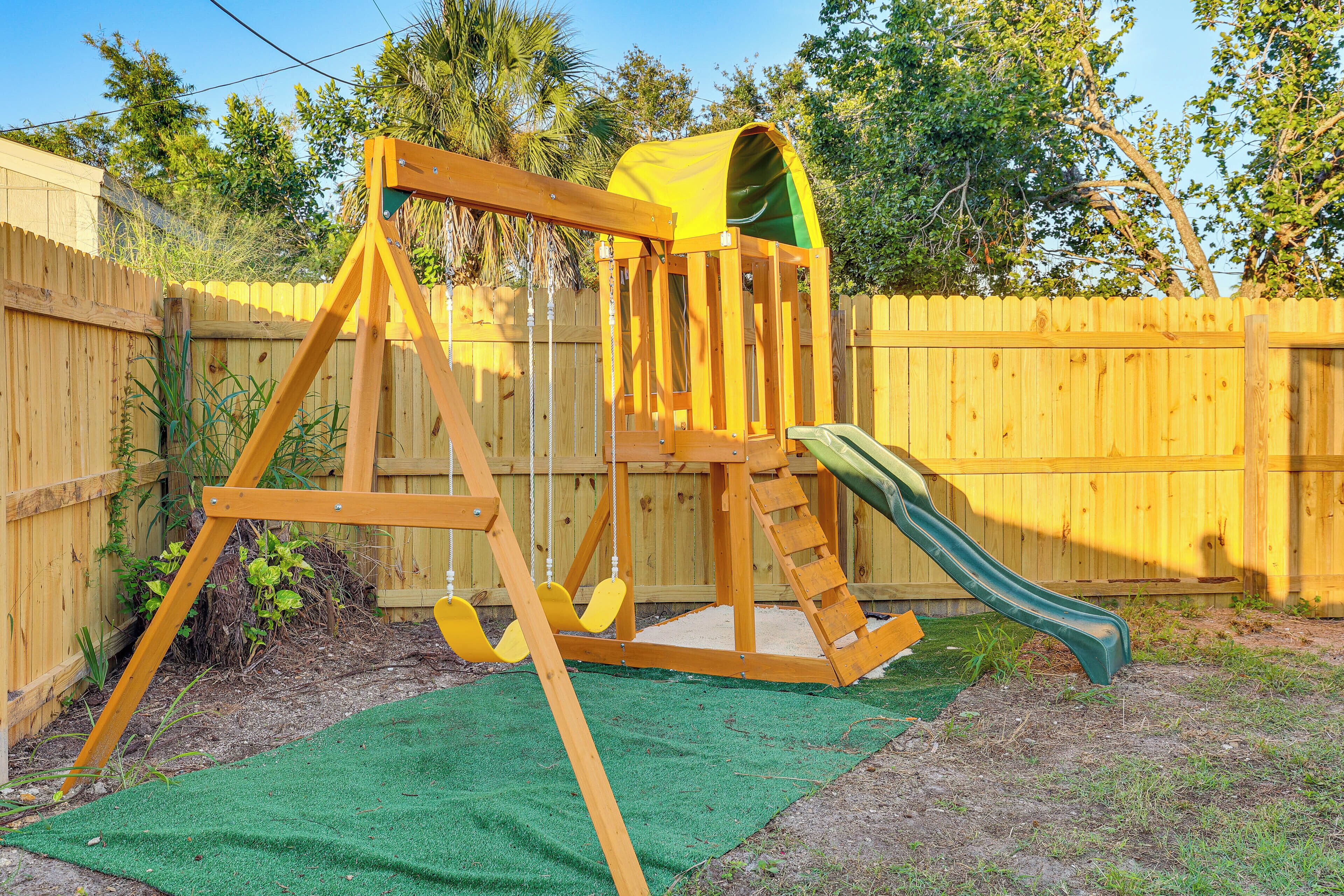 A wooden swing set with a slide, green artificial turf, and a wooden fence in the background.