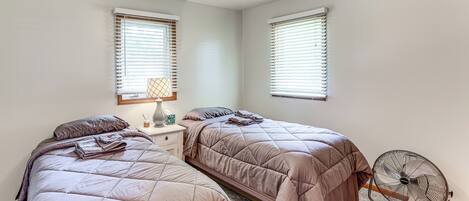 A bedroom with two beds, a bedside table with a lamp, and a standing fan.