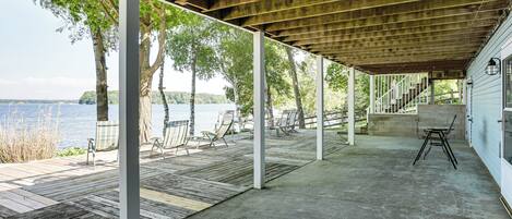 Un espace extérieur abrité, doté d’une terrasse en bois, d’un sol en béton et de piliers blancs. On y trouve des chaises et une table, et la vue porte sur une étendue d’eau et des arbres en arrière-plan.