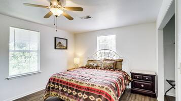 Une chambre à coucher comprenant un lit, un ventilateur de plafond, deux fenêtres, une table de chevet et une lampe fixée au mur.