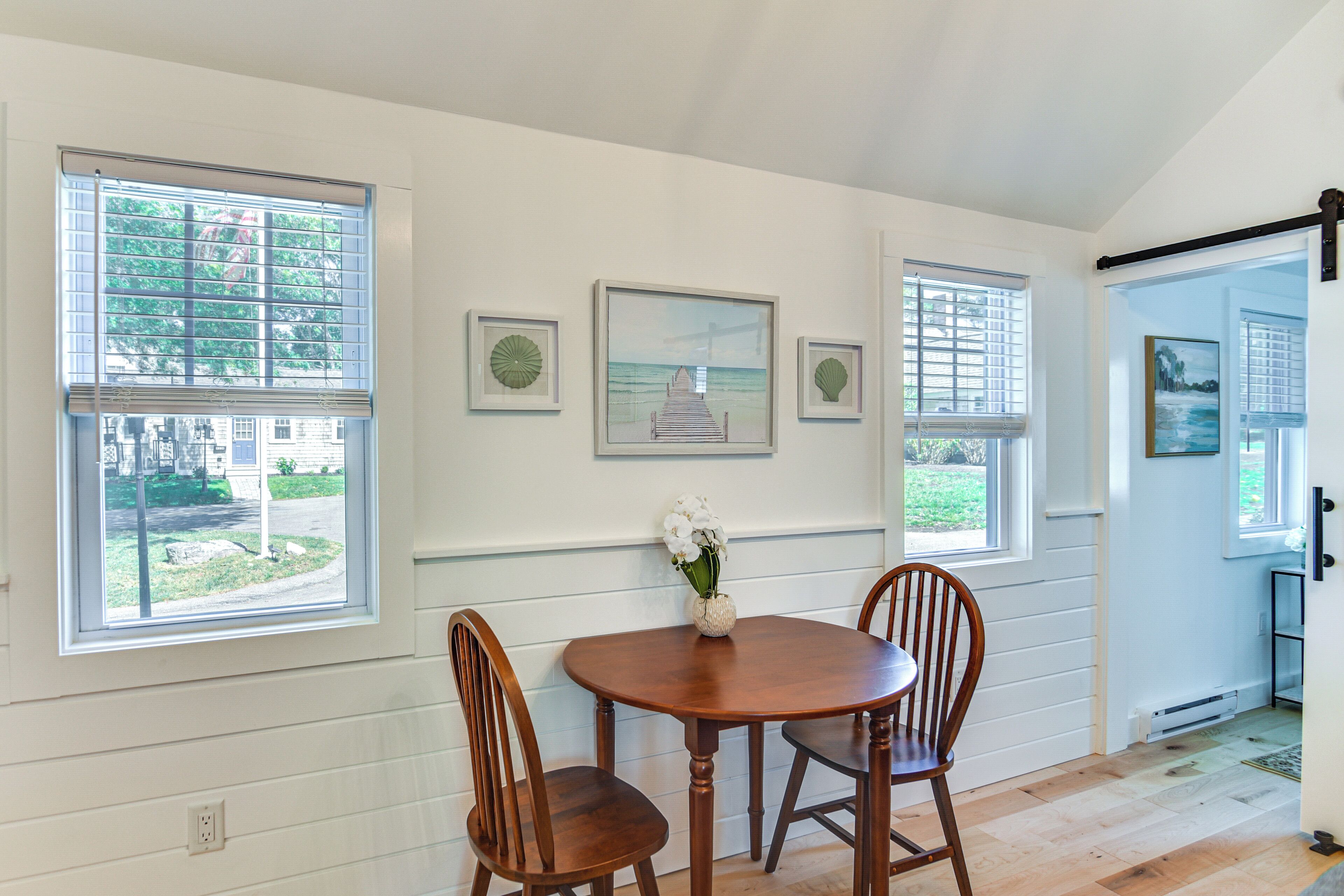 A dining area with a wooden table and chairs, framed artwork on the wall, and large windows with blinds.