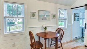 Un comedor con mesa y sillas de madera, un cuadro en la pared y ventanas grandes con persianas.