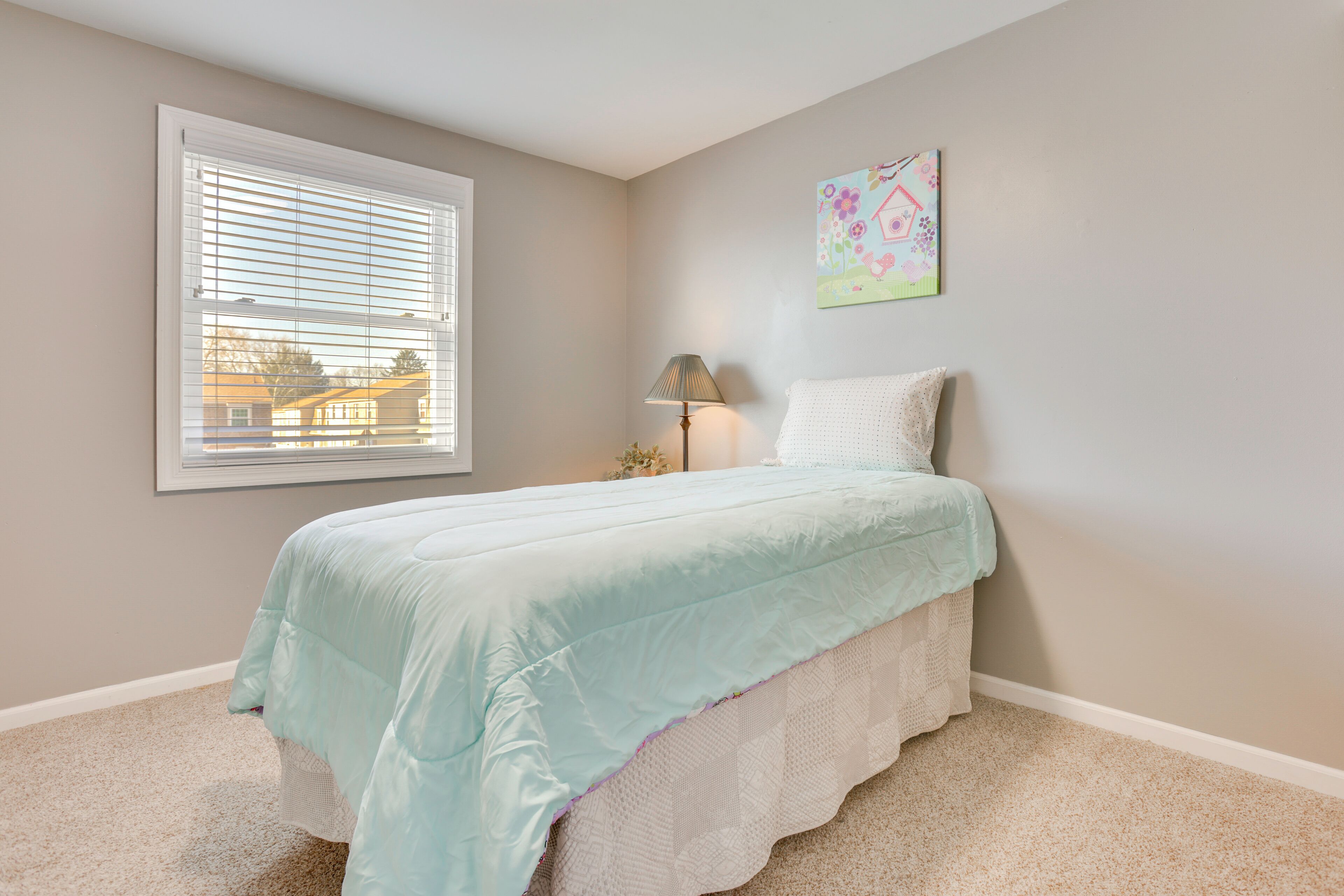 A bedroom with a bed, a window with blinds, a lamp, and a wall art piece.