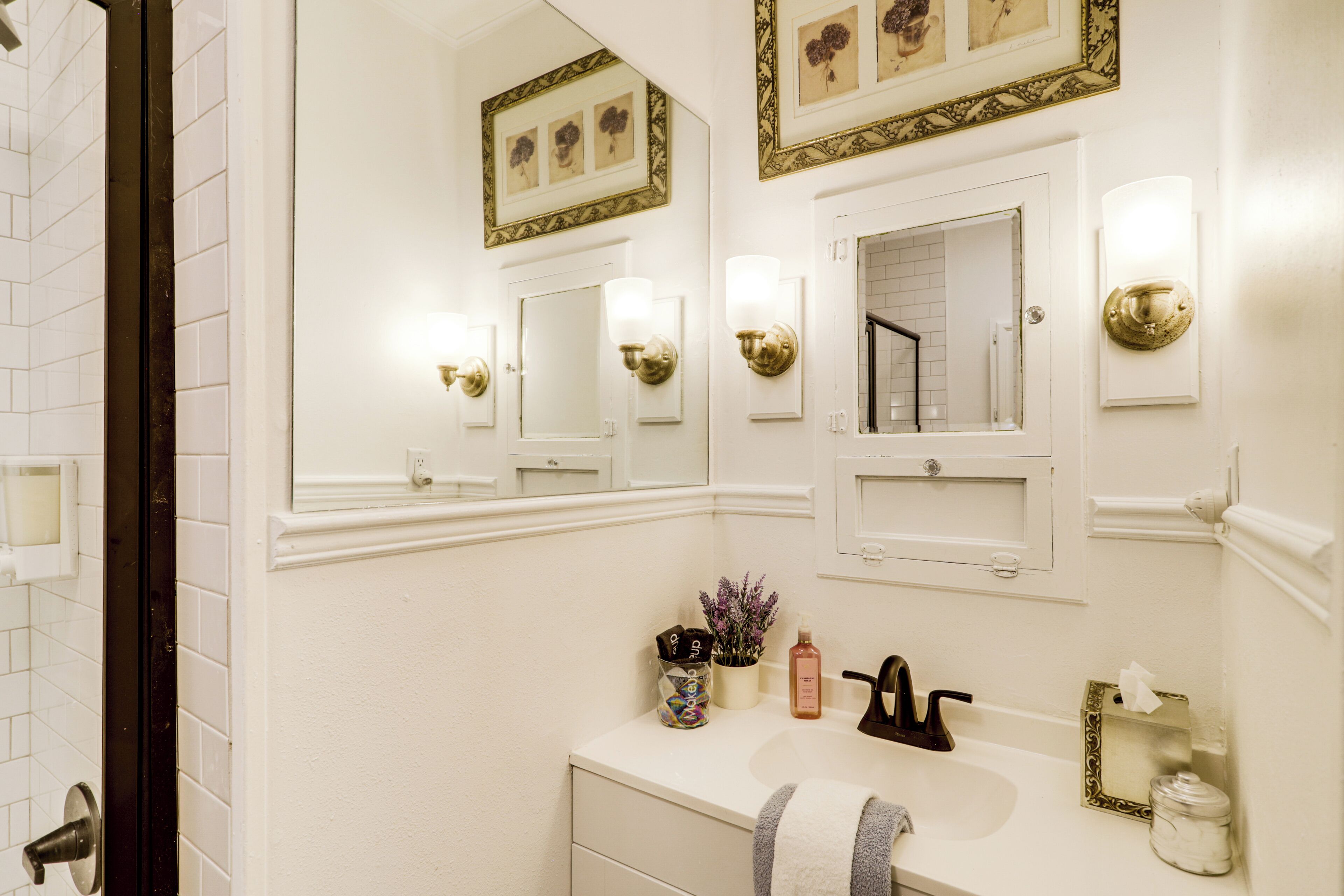 A bathroom with white walls, a white sink, and a mirror. There are framed pictures on the wall and a towel hanging on the door.