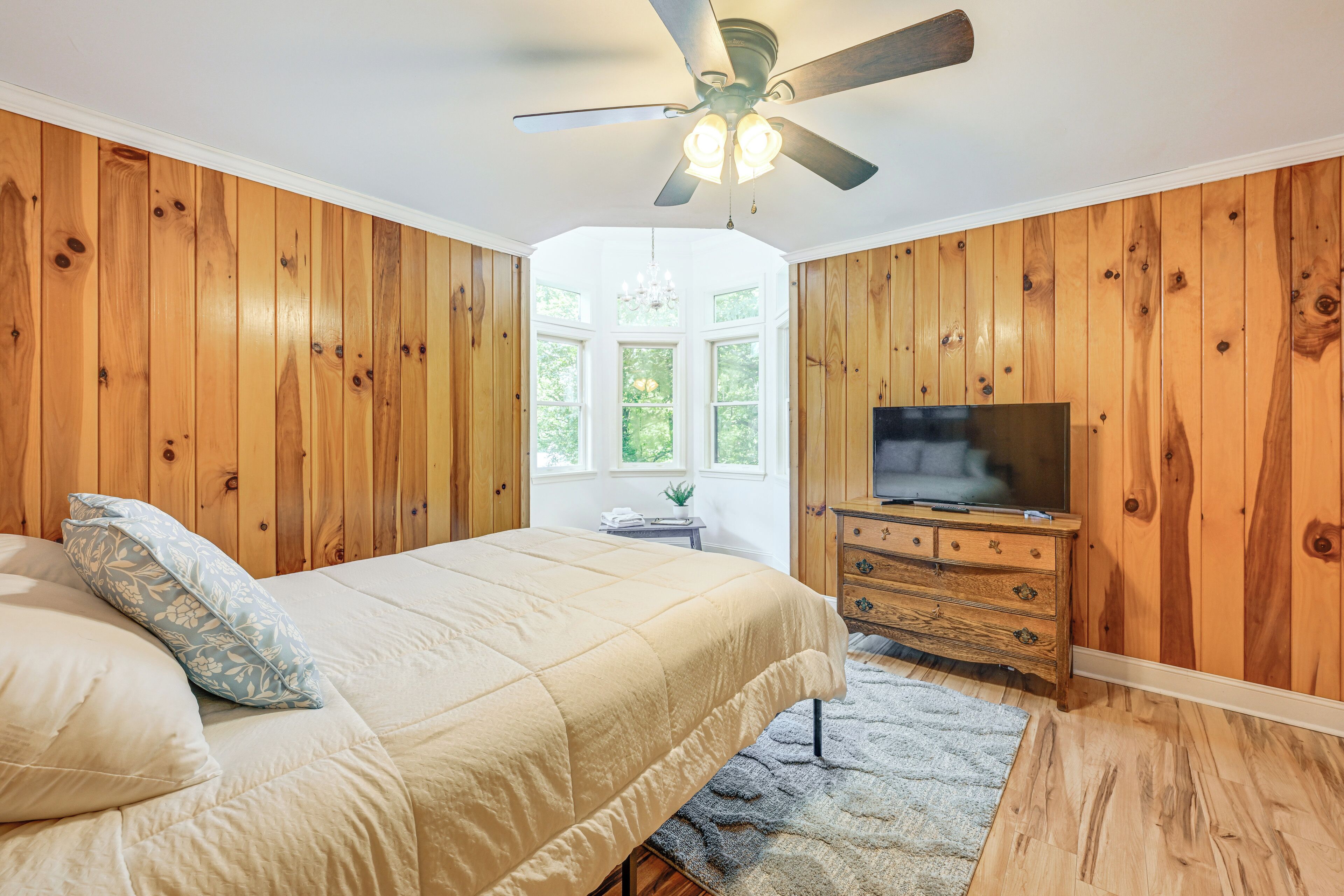 A bedroom with wooden paneling, a bed with a beige comforter, a ceiling fan, a television, and a window with a view of greenery.