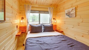 A wooden cabin bedroom with a bed, curtains, a painting, and two wall-mounted lamps.