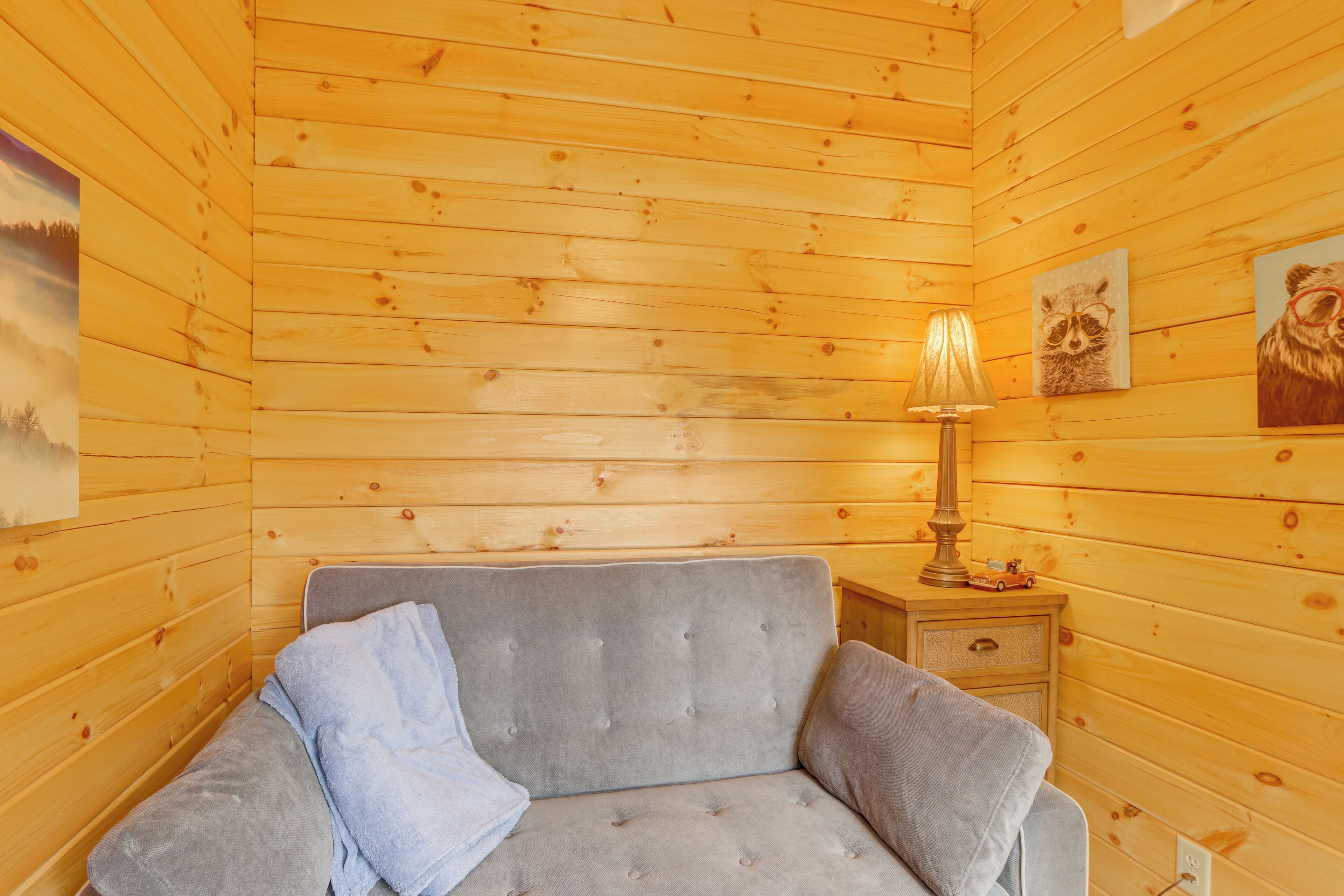 A cozy room with wooden walls, a grey sofa, a lamp, and two raccoon-themed paintings.