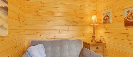 A cozy room with wooden walls, a grey sofa, a lamp, and two raccoon-themed paintings.
