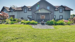 A large, modern two-story house with a symmetrical design, featuring a central entrance with a porch, multiple windows, and a well-maintained front yard with a gravel area and various plants. - Grill Out, Play Pool: Huge Home on Lake Champlain! (Chazy)