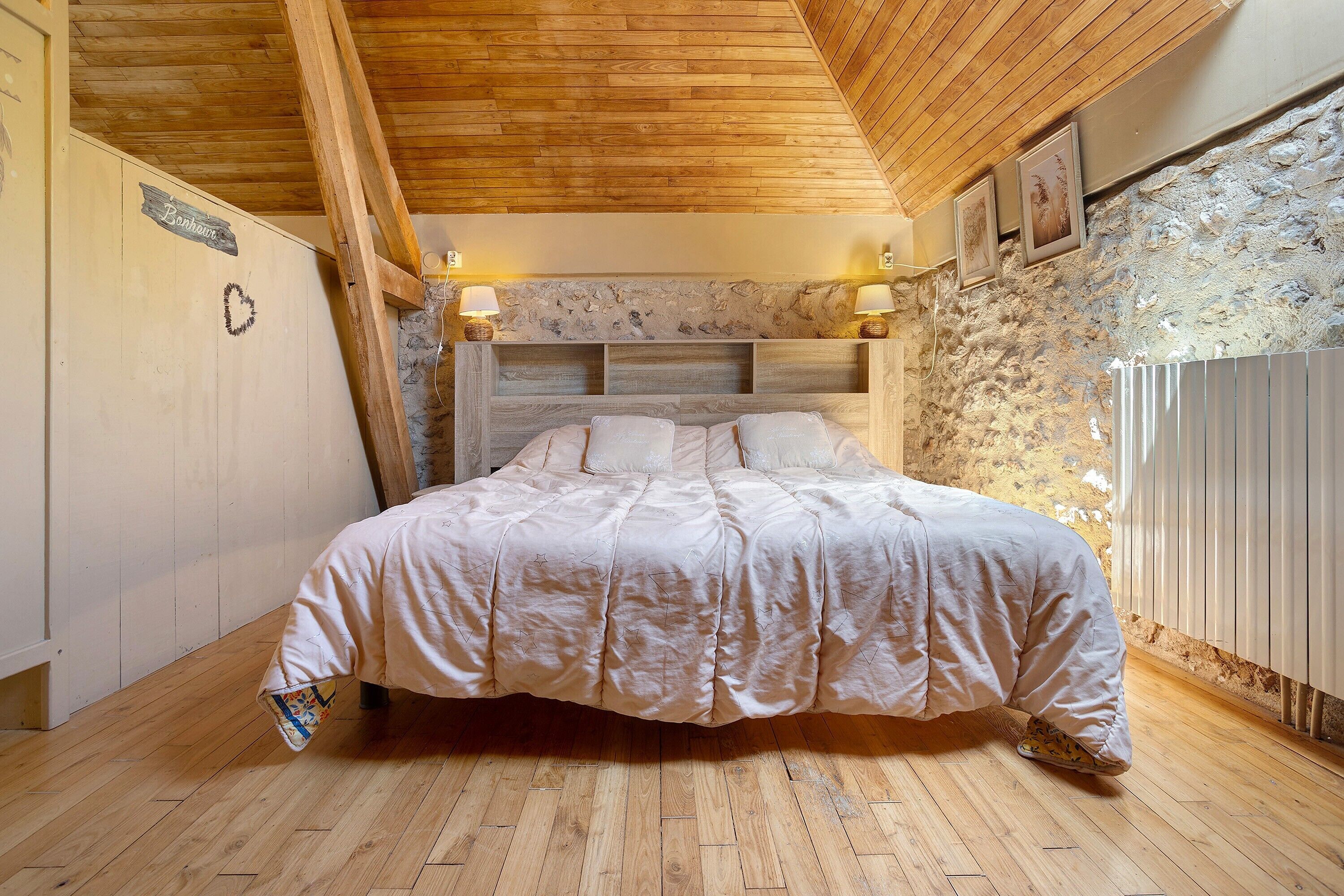 Un dormitorio con techo de madera, pared de piedra, cama con cabecera y dos lámparas fijadas a la pared.