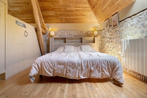 Un dormitorio con techo de madera, pared de piedra, cama con cabecera y dos lámparas fijadas a la pared.