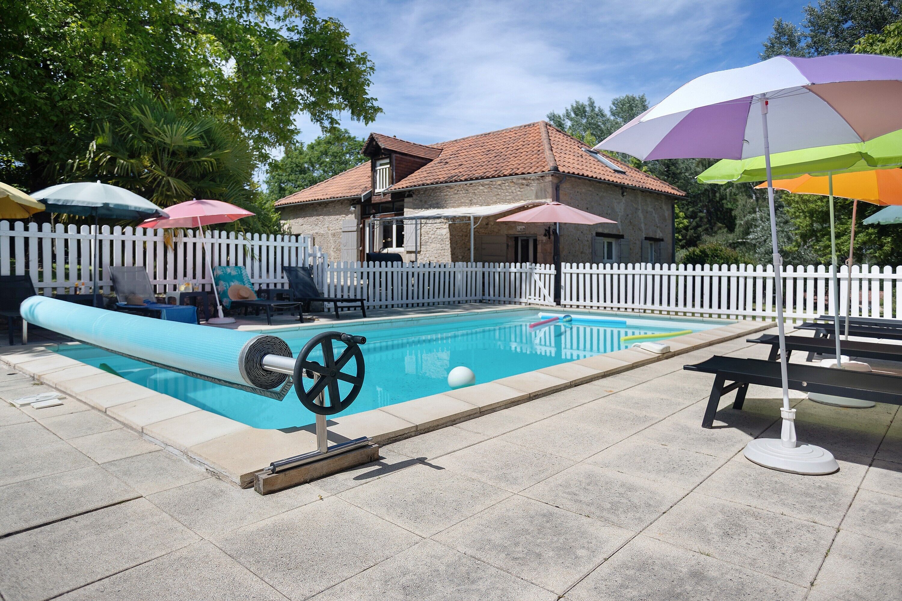 A swimming pool with a white fence, a house in the background, and multiple umbrellas.