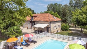A swimming pool with lounge chairs and umbrellas, a white picket fence, and a stone building with a tiled roof.