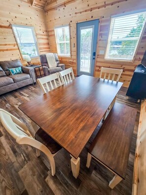 Interior de una cabaña de madera con una mesa de comedor, sillas, un sofá y una chimenea.